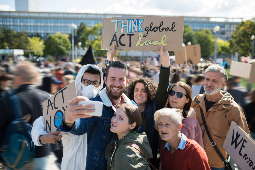 A group of six people holding signs at a protest, gathering to be included in a selfie taken with a phone