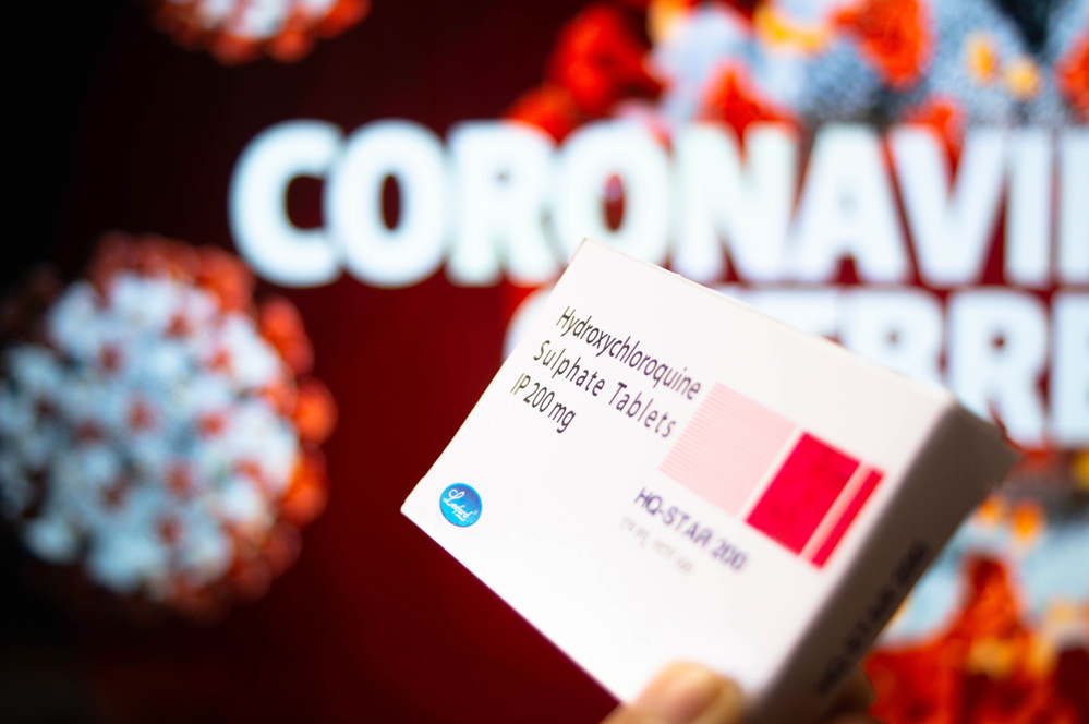A box of hydroxychloroquine sulphate tablets held by a hand with coronavirus written in background