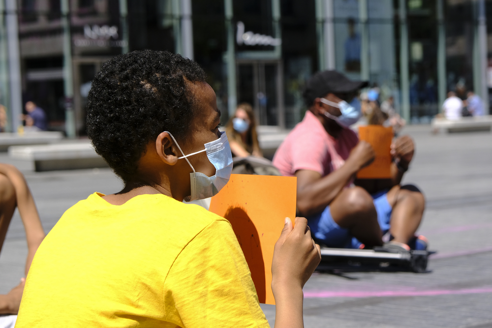 Young person sitting on cement wearing a mask and holding a sign, turned away from camera. More people also sitting and holding signs are visible in the background.