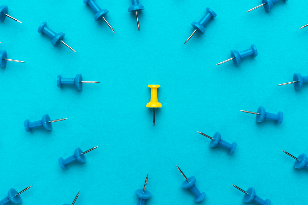 photograph of yellow push pin in the center of blue push pins with their spike turned to the yellow one