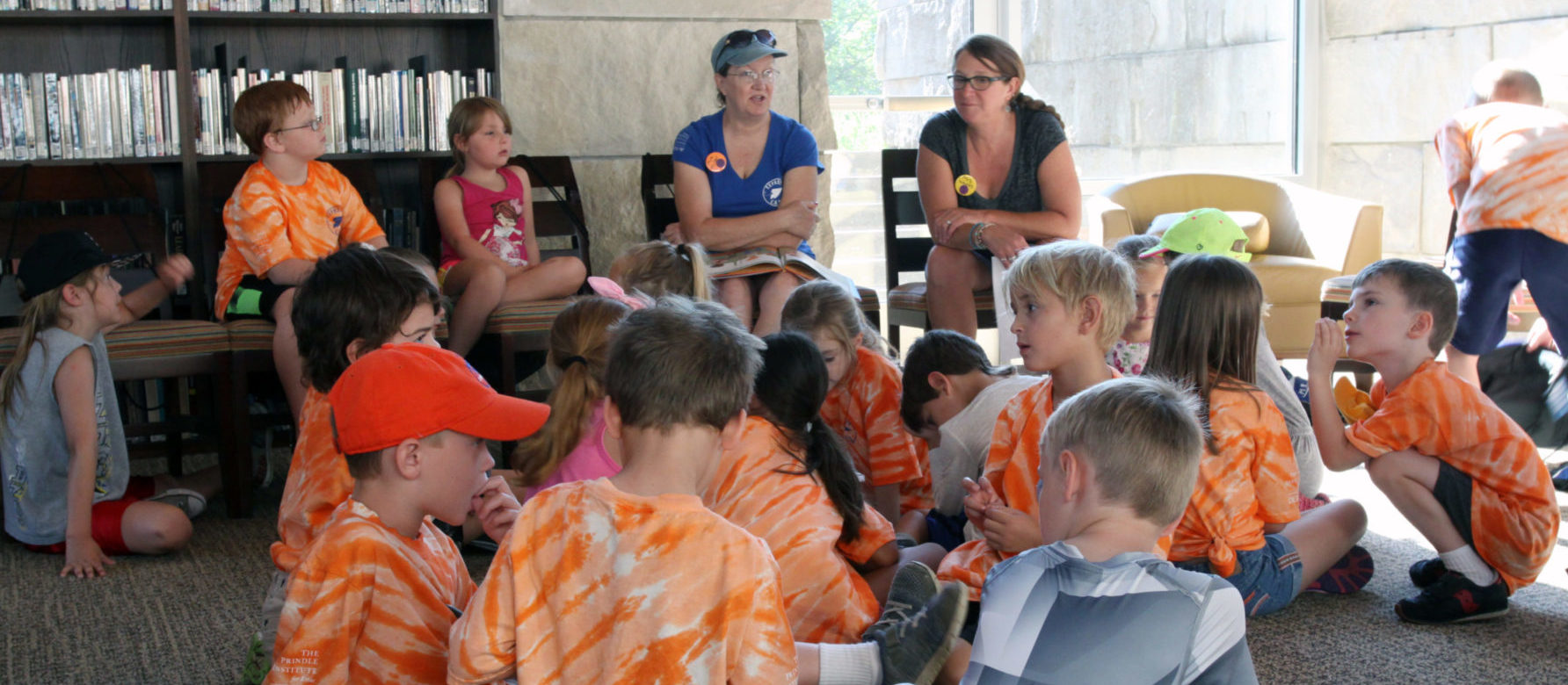 Large group of young children dressed in summer clothing gather in a semicircle on the floor around two white adult women who appear to be speaking in an animated fashion