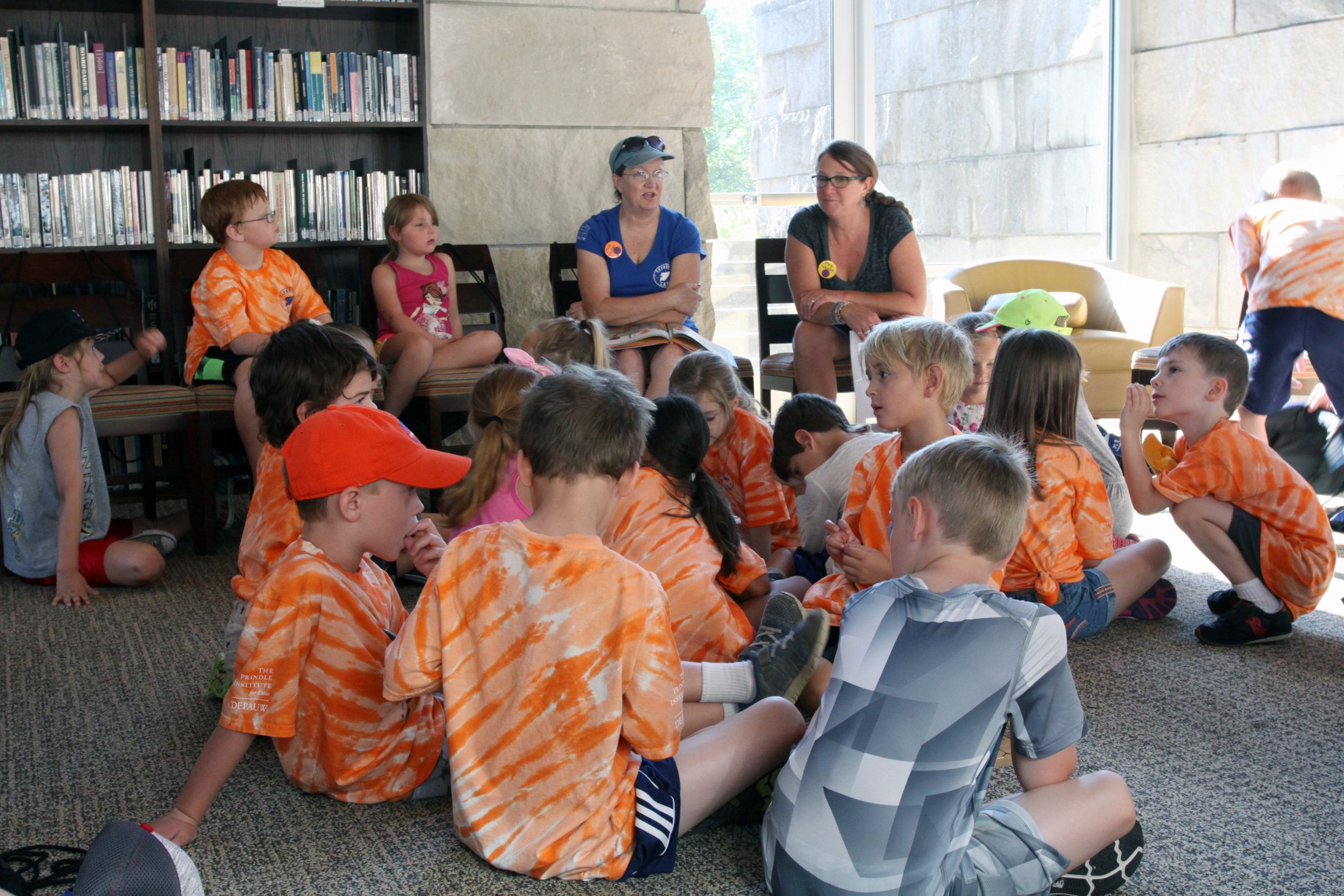 Large group of young children dressed in summer clothing gather in a semicircle on the floor around two white adult women who appear to be speaking in an animated fashion