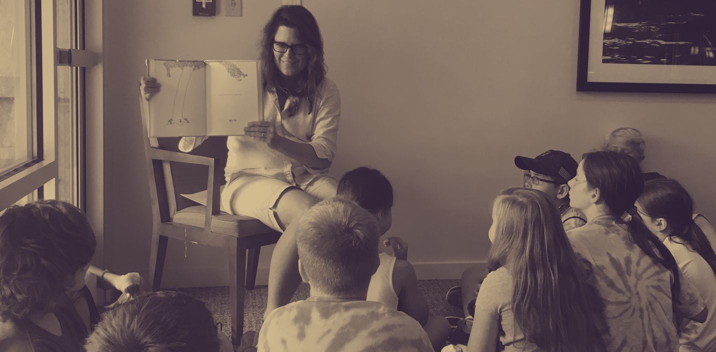 Sepia-toned photograph of a white woman holding a book open in her hands and smiling down at a group of children gathered around her on the floor to listen to the book she is reading