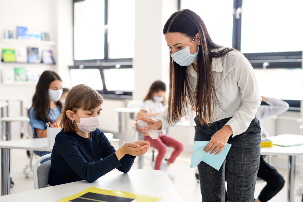 photograph of school children with face masks having hands disinfected by teacher