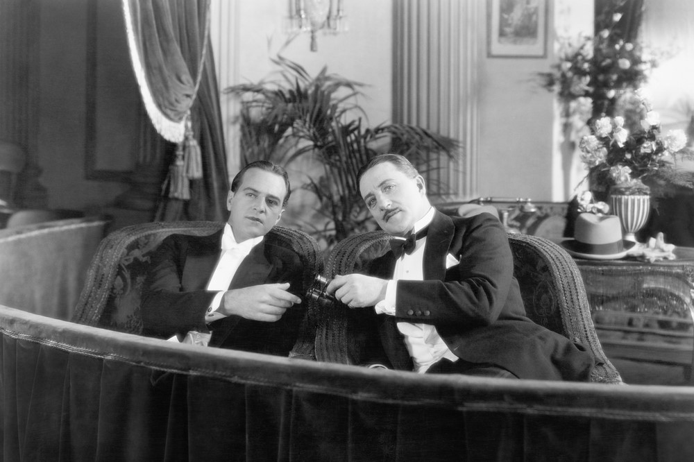 black-and-white photograph of two white men sharing opera glasses ina theater box