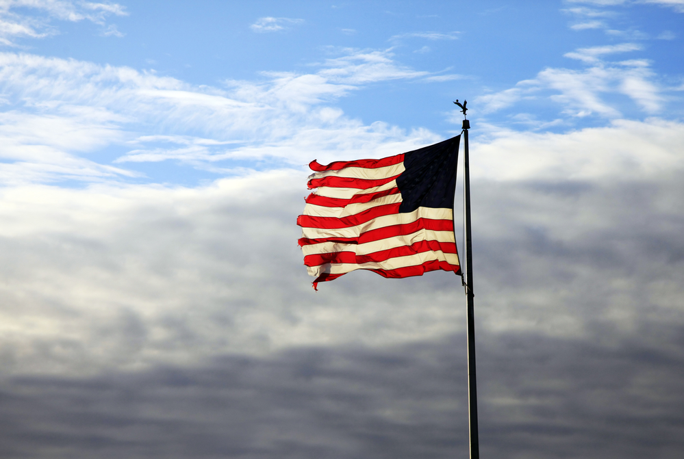 photograph of worn USA flag on pole with clouds behind