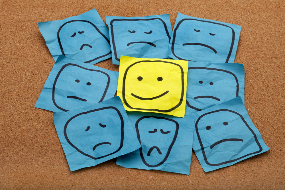 photograph of happy smiley face on yellow sticky note surrounded by sad unhappy blue faces