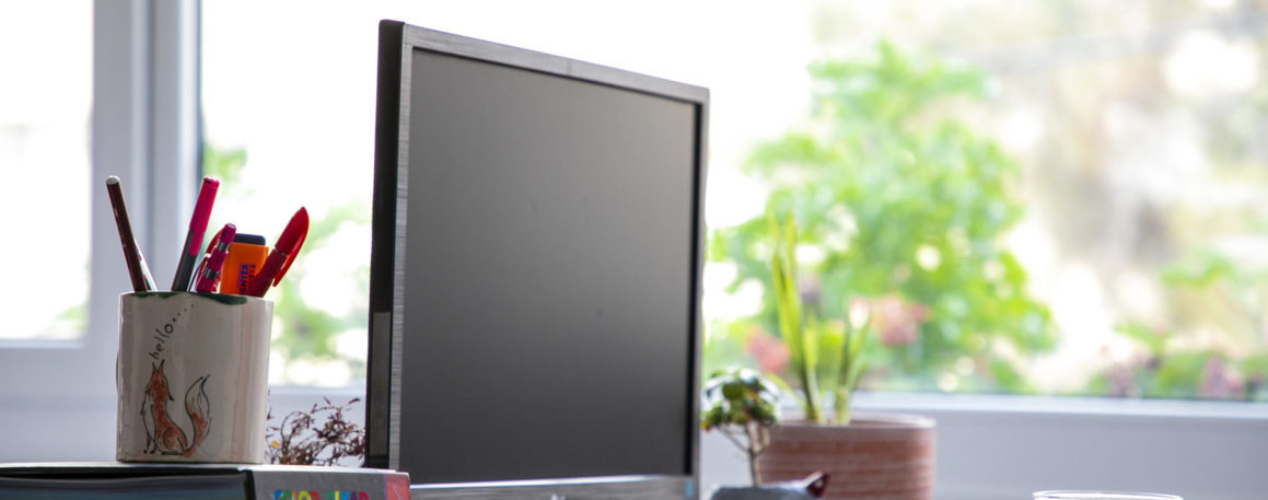 Photograph of a computer monitor, cup of tea, stack of books and potted plant sitting on a desk in front of a blurry window.