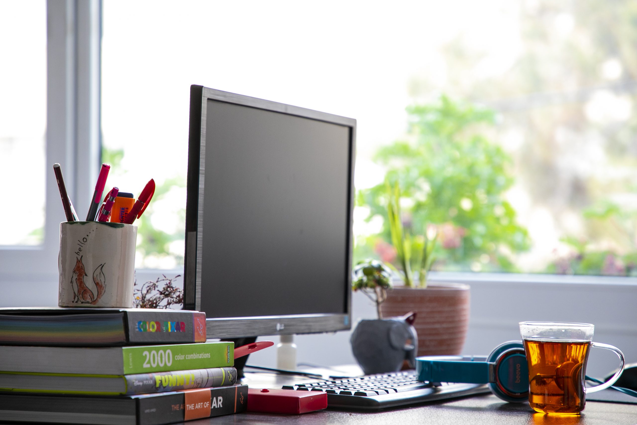 Photograph of a computer monitor, cup of tea, stack of books and potted plant sitting on a desk in front of a blurry window.