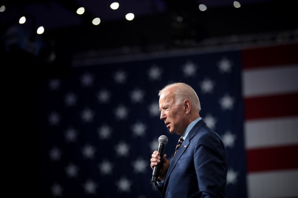 photograph of Joe Biden speaking with microphone with American flag in background