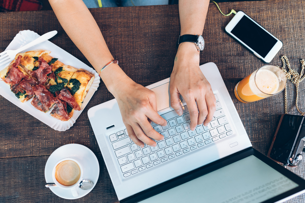 overhead photograph of hands on laptop on dinner table surrounded by food and beverages