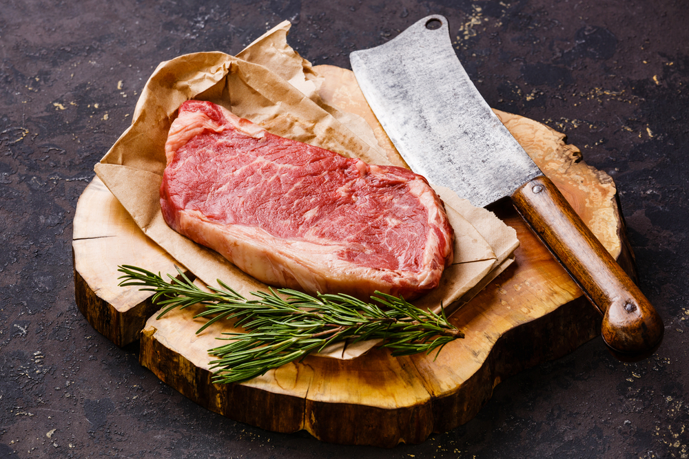 photograph of raw steak arranged on butcher block with cleaver and greenery