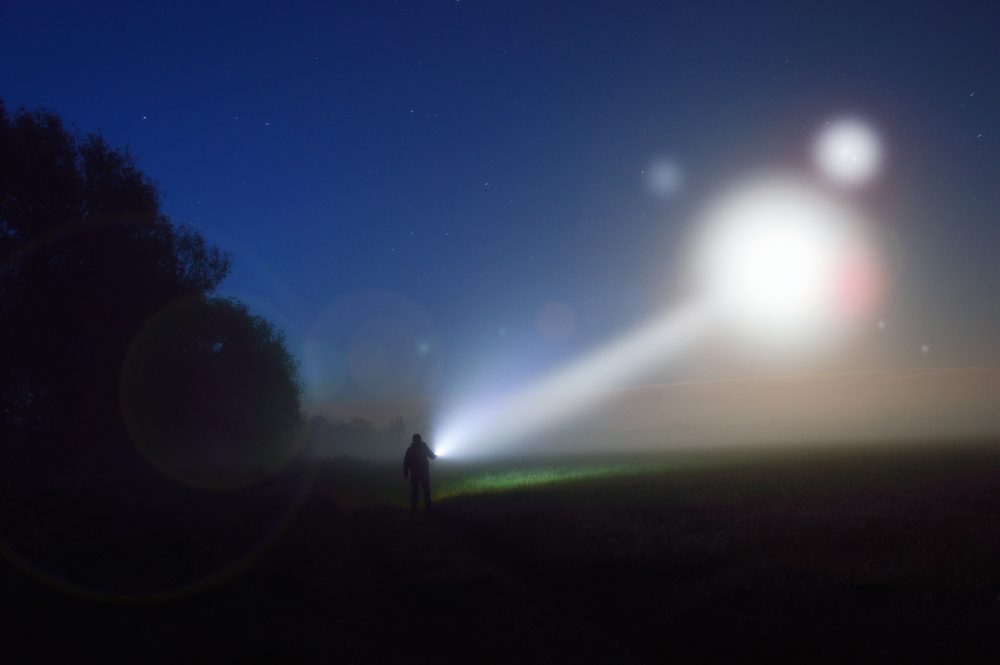 photograph of silhouetted figure shining flashlight at light source in the night sky