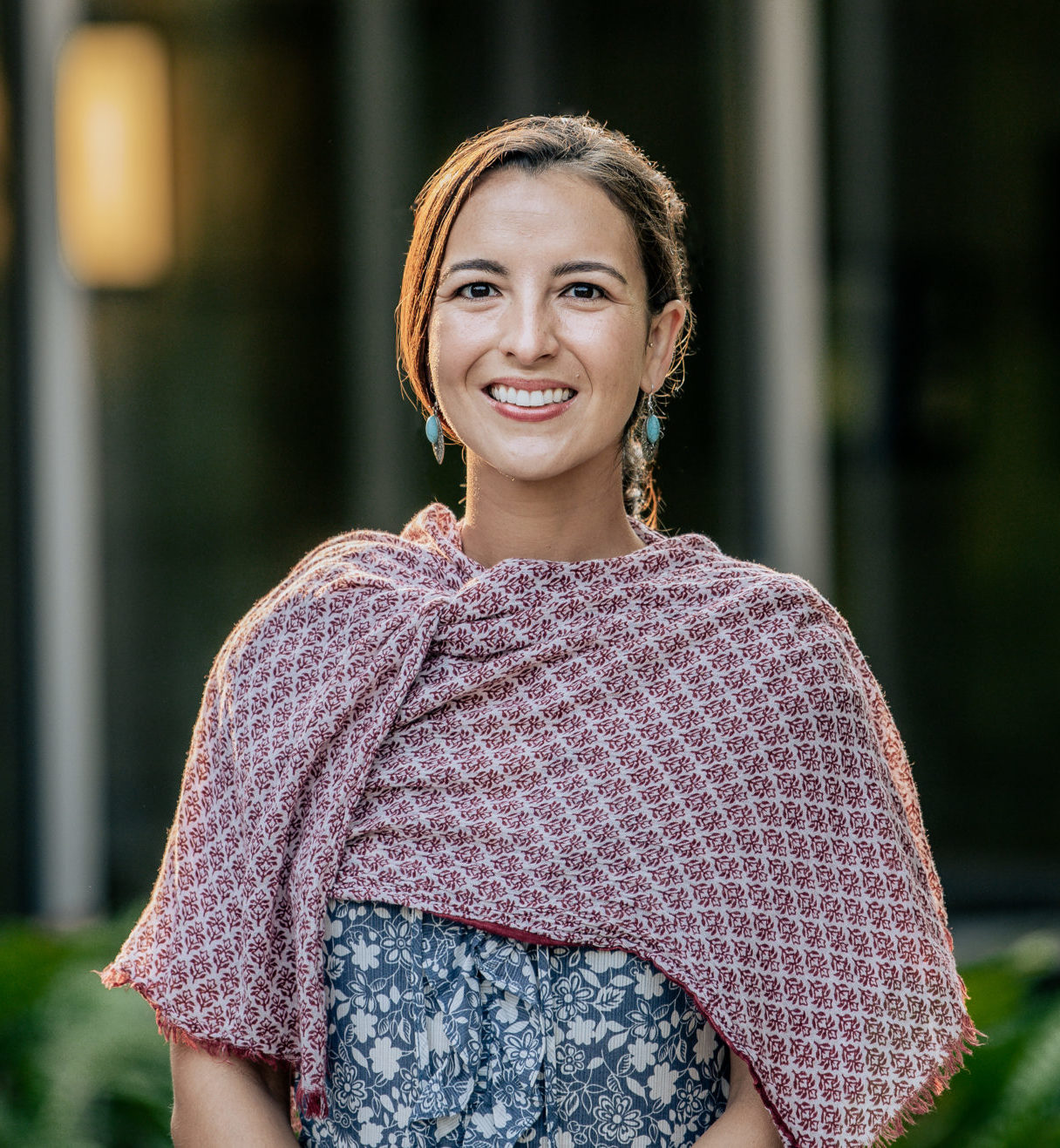 Color photograph of a woman with a red and white shawl over her shoulders. She is smiling and standing in front of a group of glass windows and greenery.
