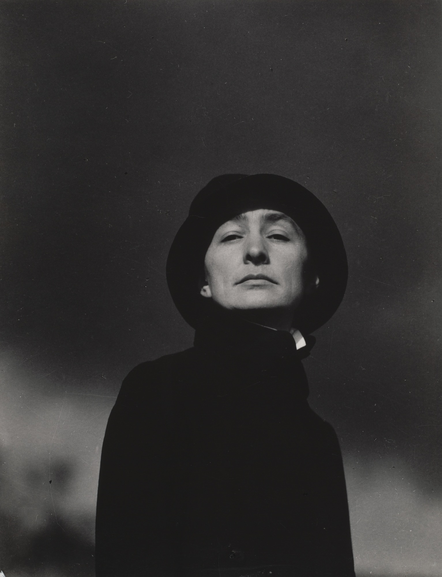 Black and white photograph of the artist Georgia O'Keeffe by Alfred Stieglitz. The photograph is taken from below, so that O'Keeffe is looking slightly down at the camera/viewer. She is wearing an all-black garment that covers her neck and arms completely. She also wears a small black hat that creates a circle around her face. She is not smiling.