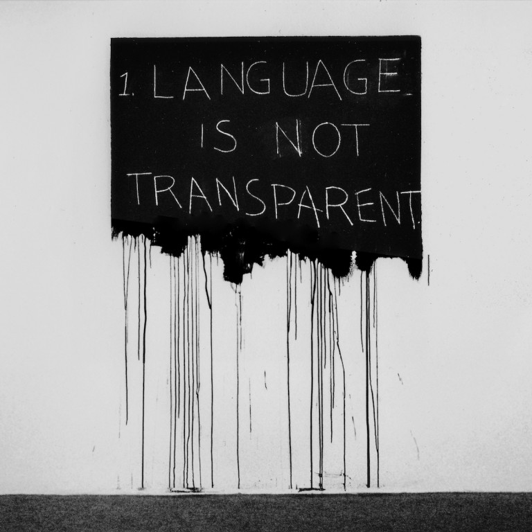 Photograph of a gallery installation of "Language Is Not Transparent" by Mel Bochner. It is a black rectangle painted directly onto a white wall. The bottom edge of the rectangle is not a straight line; it is uneven and has many drips of black paint that run almost to the gallery floor. The following handwritten message is written in white chalk in all capital letters inside the black rectangle: "1. LANGUAGE IS NOT TRANSPARENT."