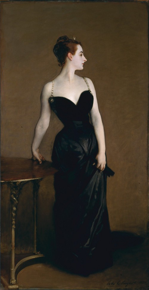 Full-length portrait of a white woman with reddish-brown hair standing in front of a table in an elegant black dress with silver straps. Her arms and neck are exposed.