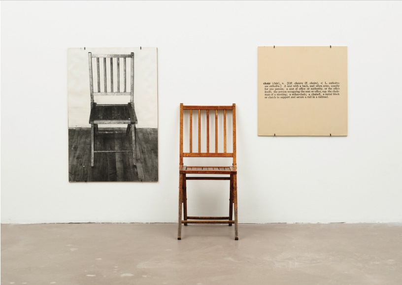 Photograph of a gallery installation of the conceptual work of art "One and Three Chairs" by Joseph Kosuth. The installation consists of a wood folding chair, a mounted photograph of a chair, and mounted photographic enlargement of the dictionary definition of the word "chair."