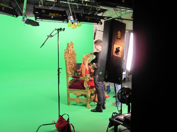 Color photograph of reality star Paris Hilton sitting on a throne in front of a green screen while many cameras point at her.