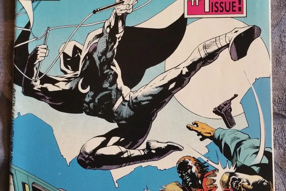 photograph of 'Moon Knight' comic cover featuring an illustration of a superhero in a jump pose with a black suit and cape
