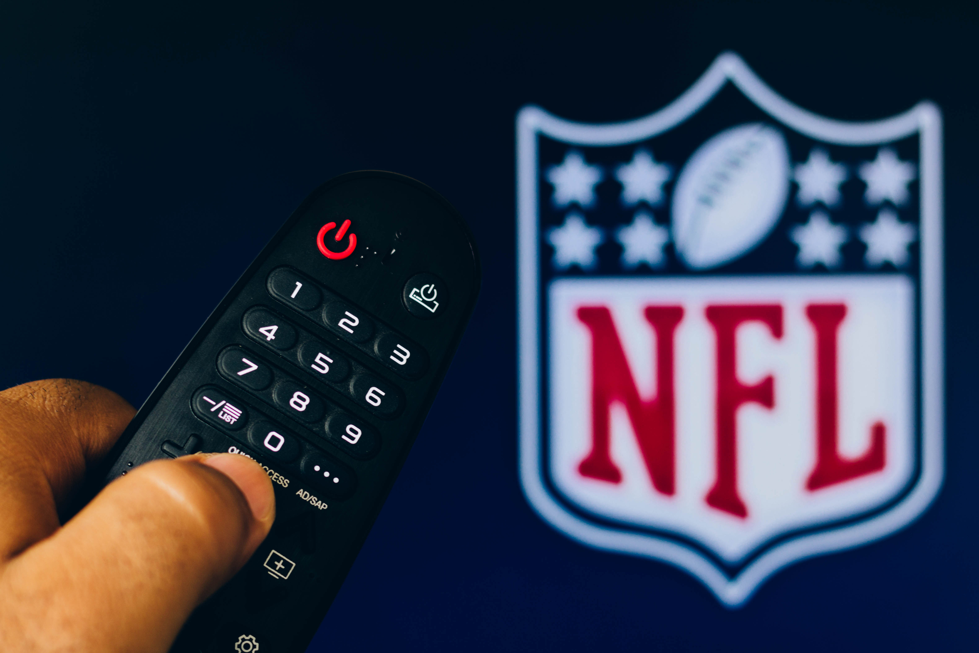 photograph of remote in hand with TV in background displaying NFL logo on screen