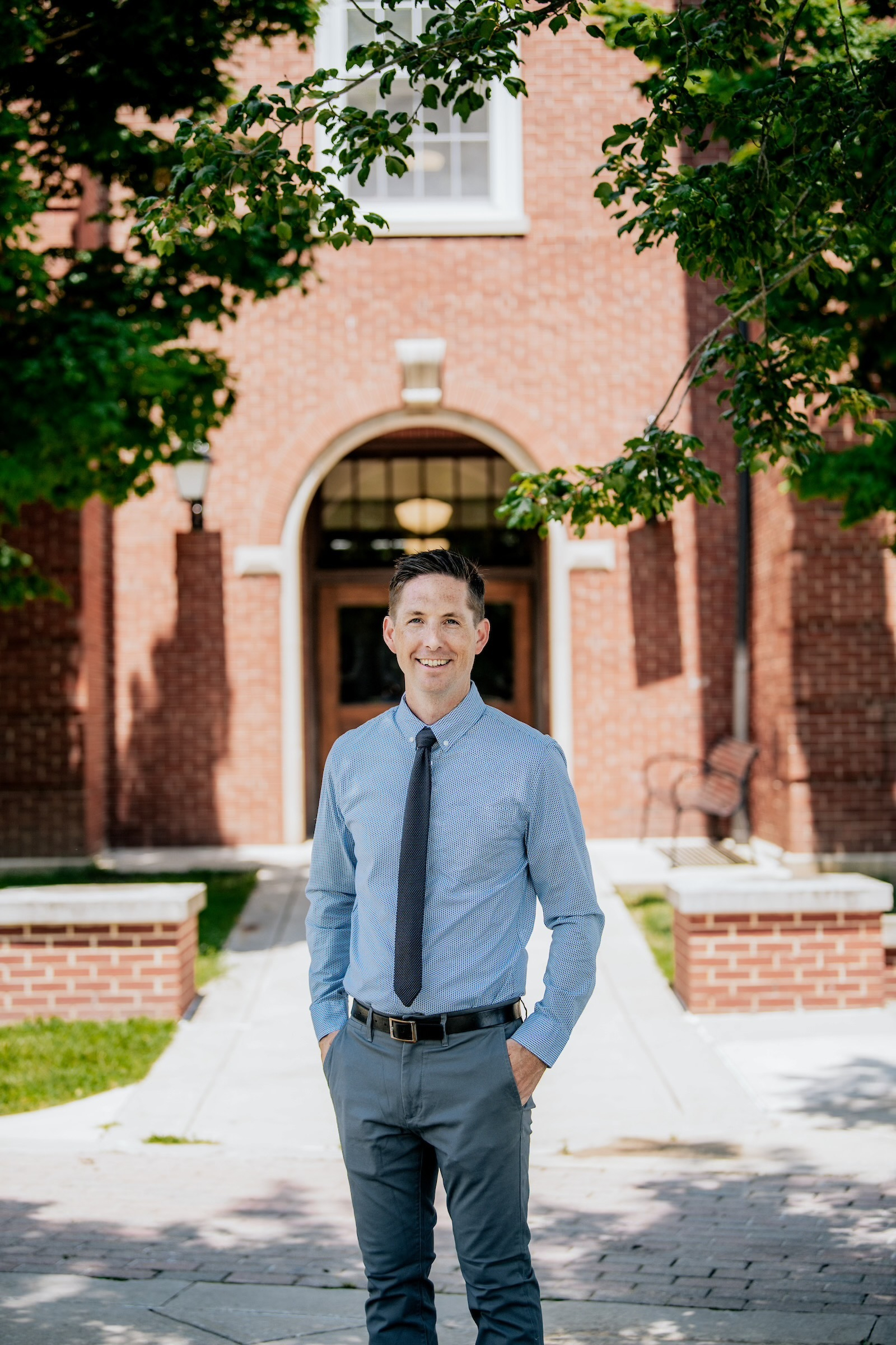 Color photograph of the Prindle Institute's executive director Jeff Dunn, a white man in a blue dress shirt and gray slacks. He is standing in front of a brick archway on the campus of DePauw University.