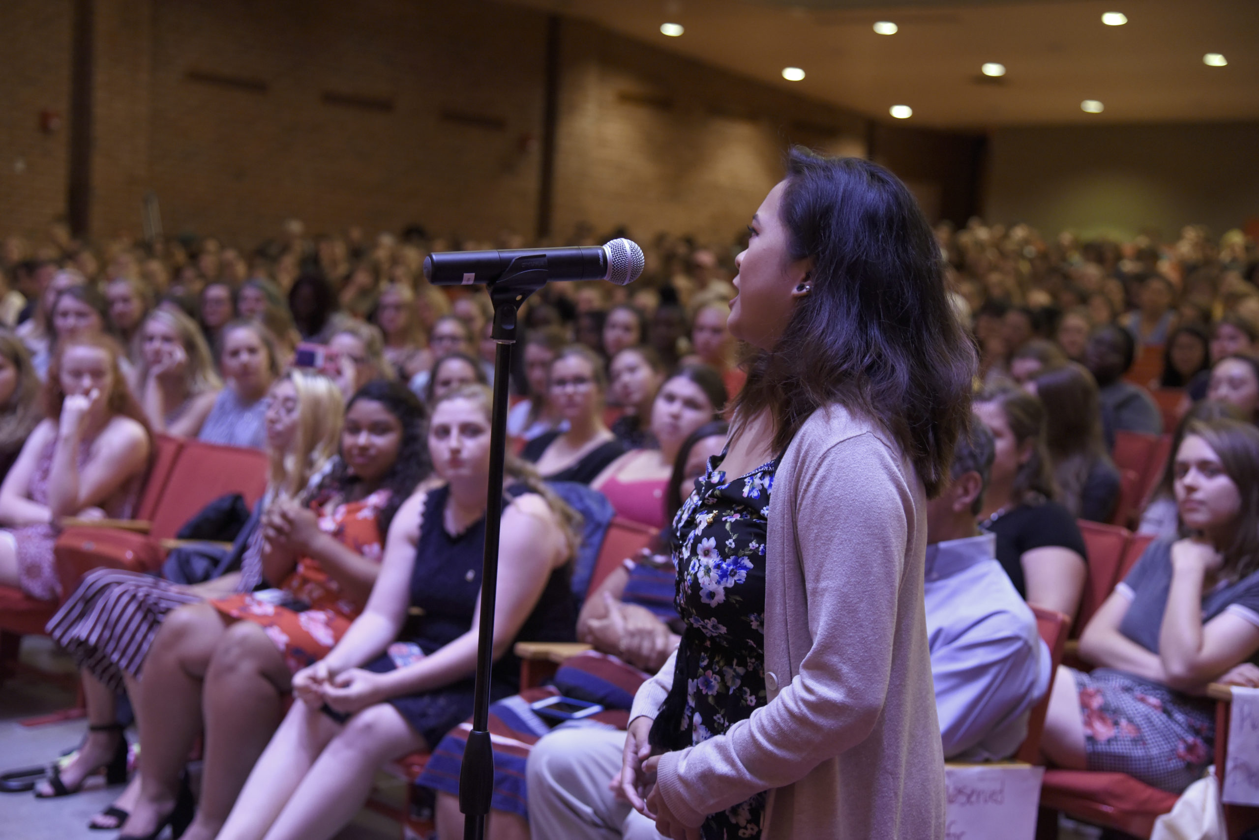 Color photograph of a crowded lecture hall with a person standing up and asking talking into a mic on a mic stand.