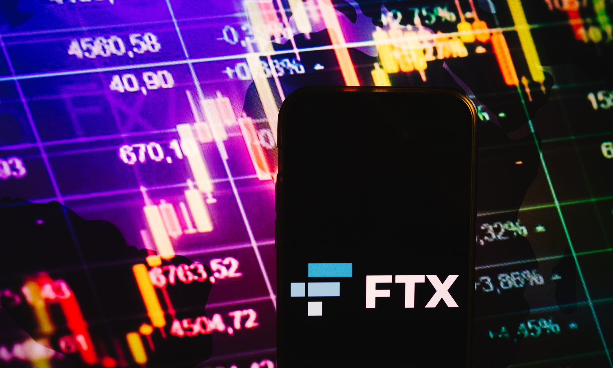 image of smartphone displaying FTX logo with stock market graphs in background