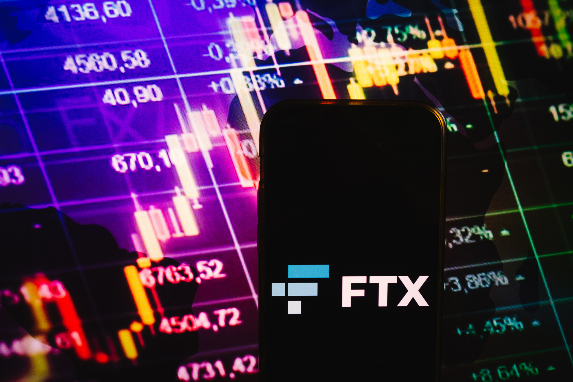 image of smartphone displaying FTX logo with stock market graphs in background