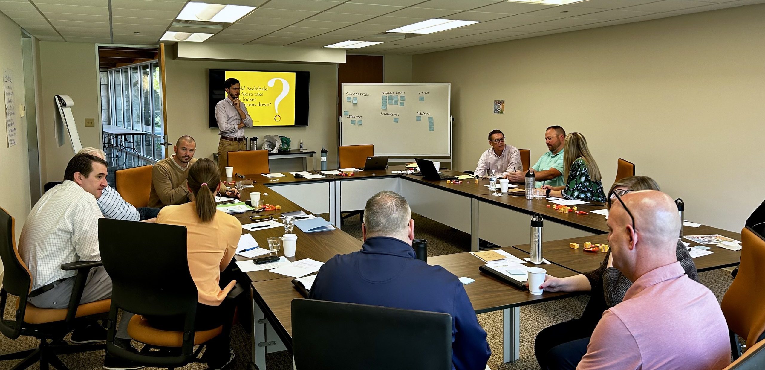 Group of adults seated at tables in a rectangular formation. A person stands at the front of the room in front of a screen and delivers an ethical reasoning workshop for decision makers.