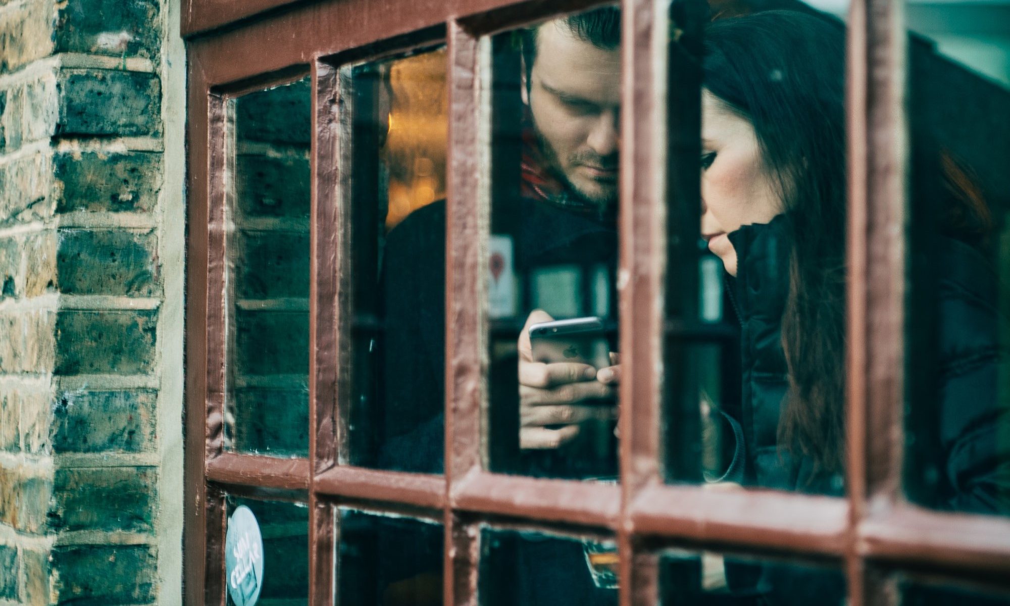 photograph of couple looking at smartphone behind window